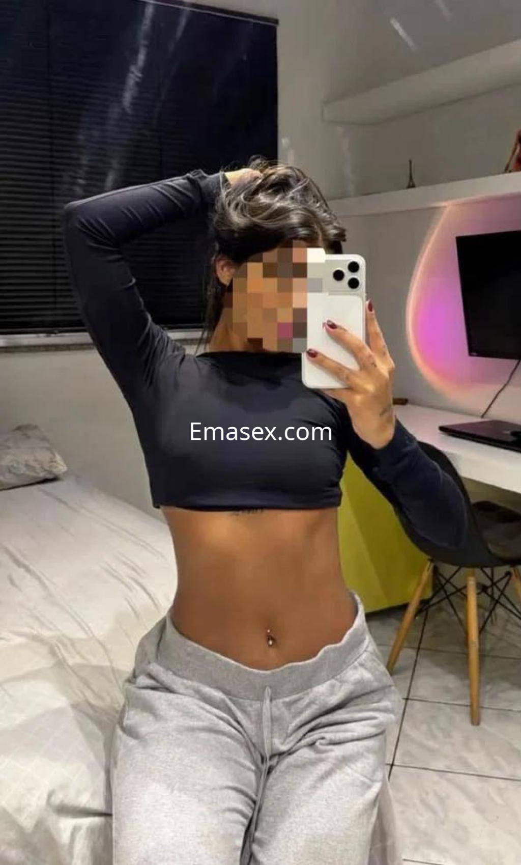 Puta en Málaga muestra su figura en un top negro y pantalones grises, con una pose relajada frente al espejo.