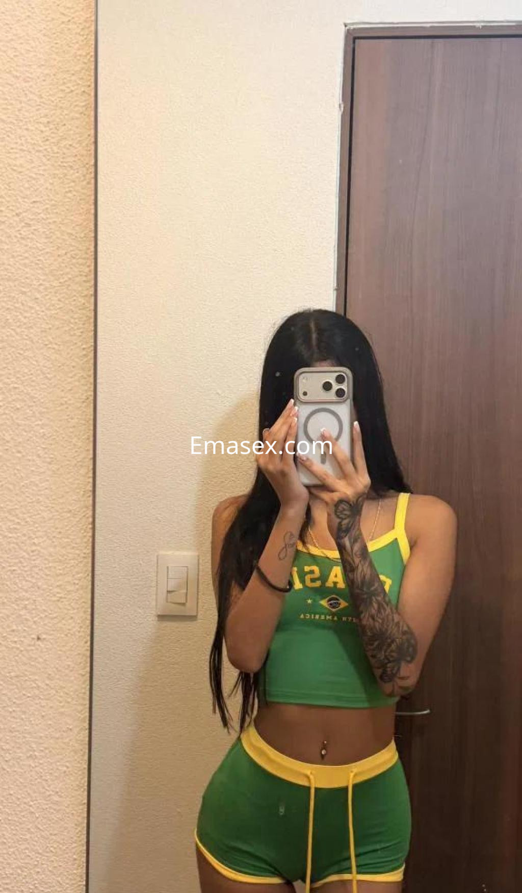 La escort en Murcia se muestra en un espejo con una camiseta verde y amarilla, luciendo un tatuaje en el brazo.