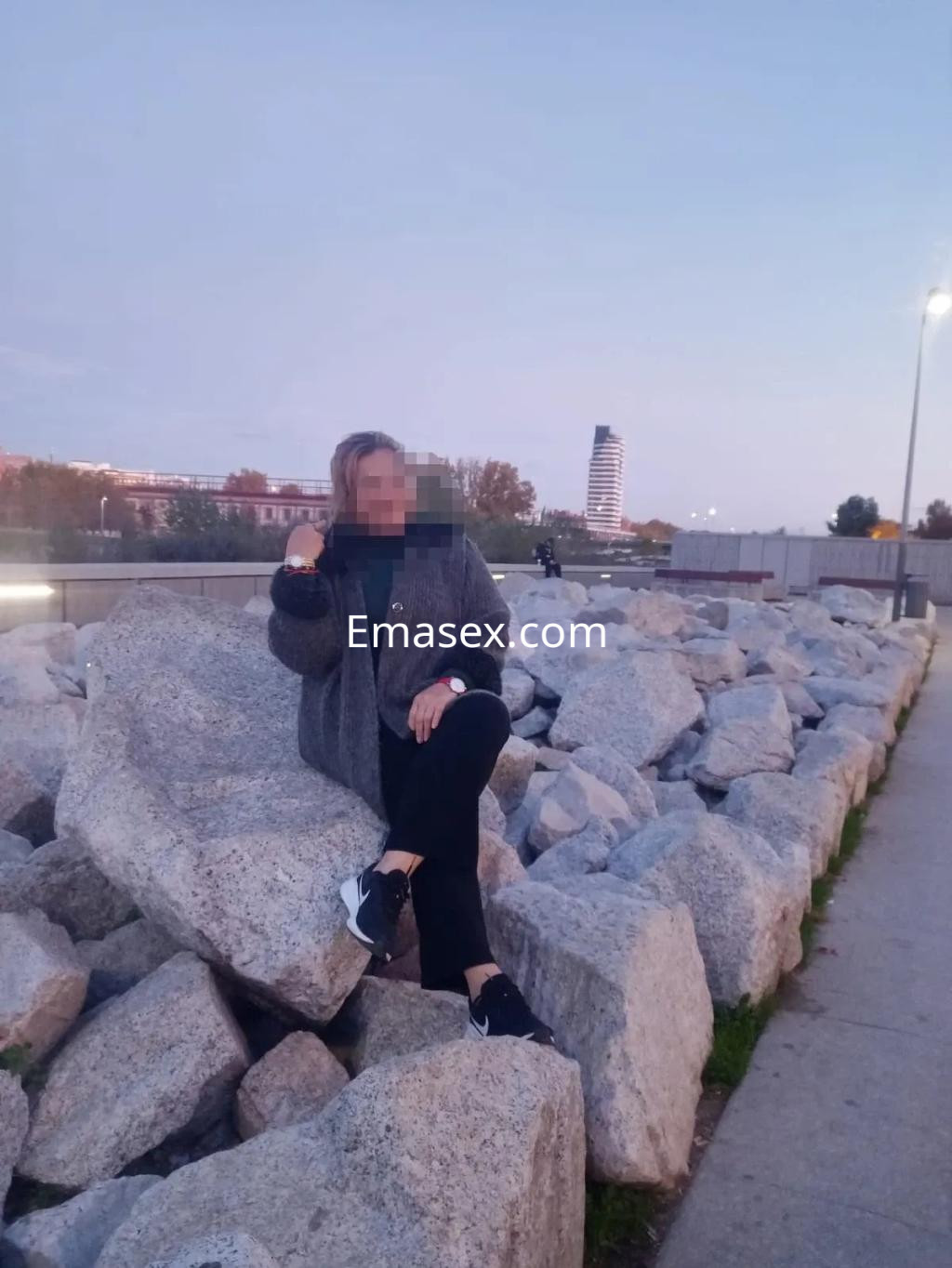 Puta sentada sobre rocas en Murcia, viste ropa cómoda y deportiva, con un suéter gris y zapatillas negras.