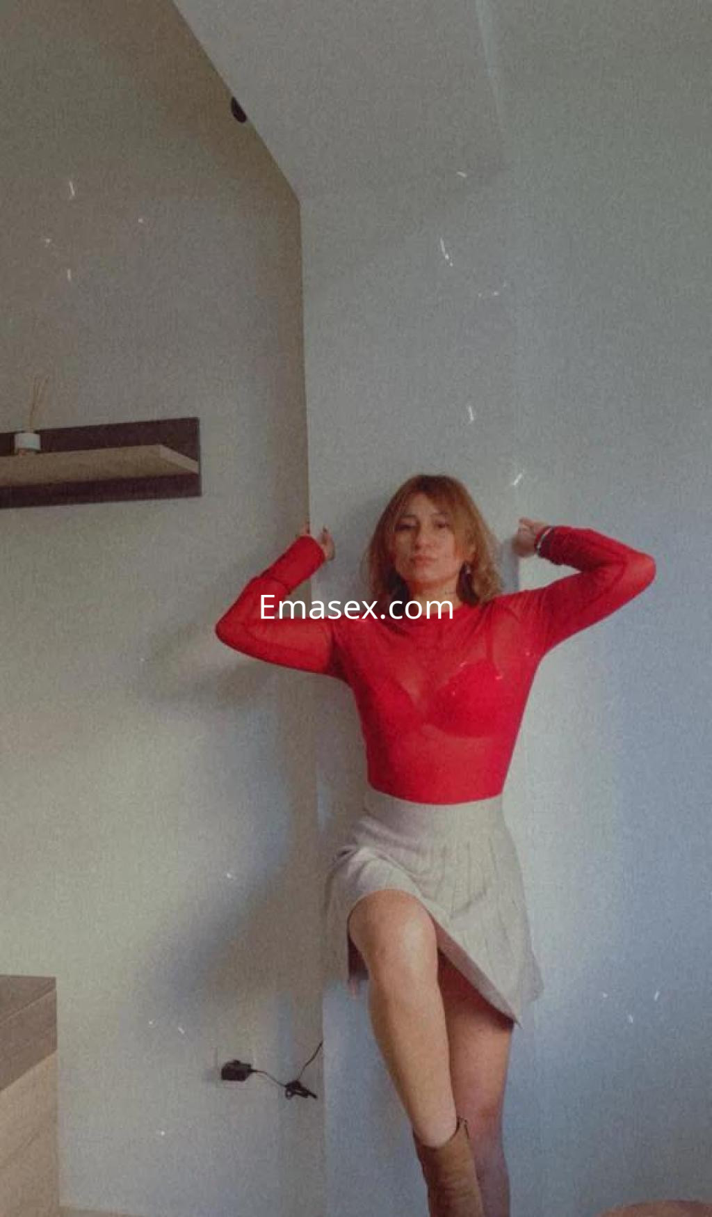Escort en Murcia con blusa roja transparente y falda corta, posando de pie con una pierna levantada.