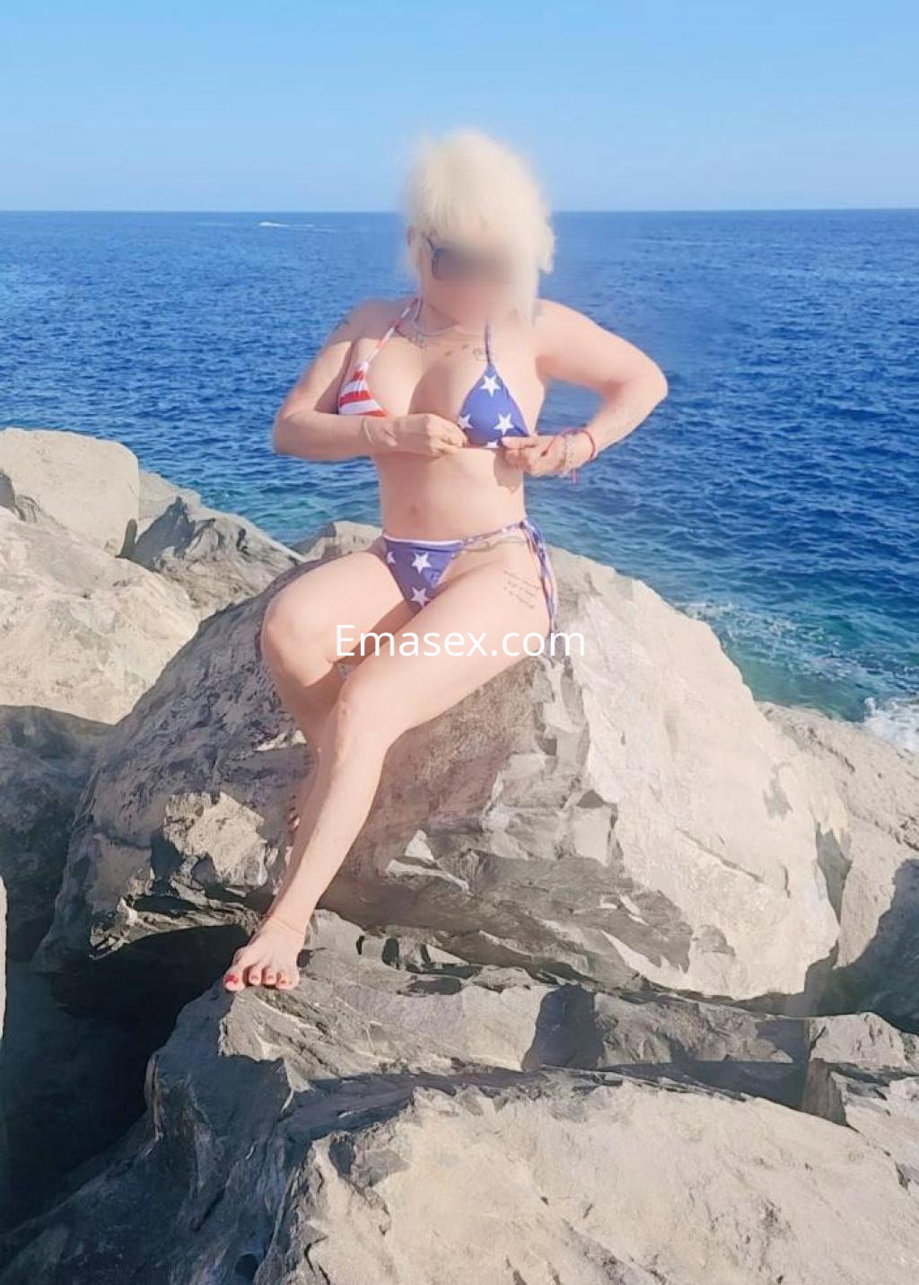 Escort en Albacete sentada sobre rocas frente al mar, vistiendo un bikini con estampado de estrellas y rayas.