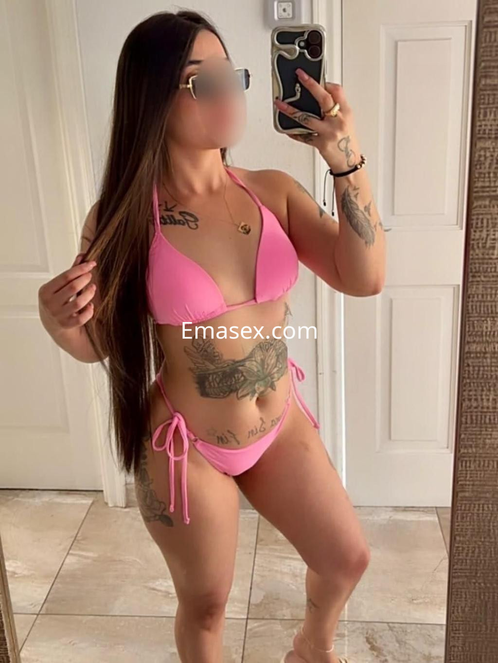 Escort en Tenerife posando frente al espejo, lleva un bikini rosa y muestra sus tatuajes y cabello largo.