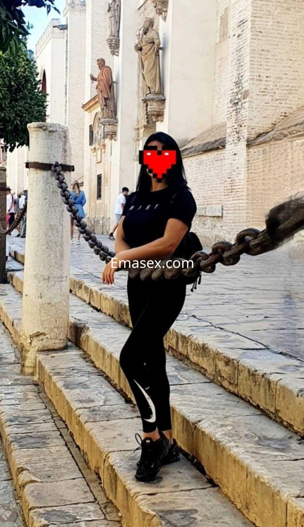 Puta en Sevilla, posando con ropa deportiva negra y zapatillas, frente a un edificio histórico.