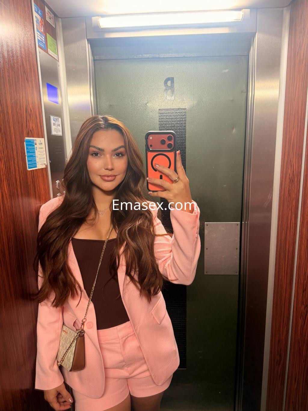 Escort en Coruña con cabello largo y suelto, vistiendo un traje rosa y posando en un ascensor con espejo.