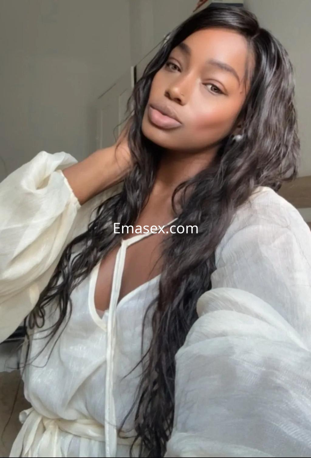 Escort en Cádiz con cabello largo y ondulado, luciendo una blusa blanca de mangas largas y un fondo luminoso.