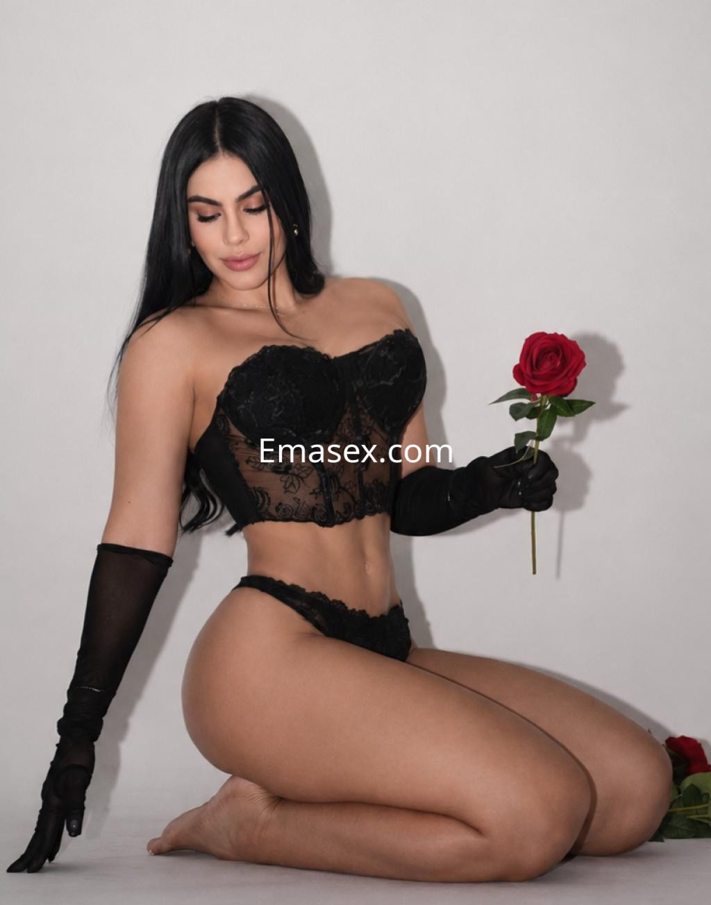 Escort en Bilbao se arrodilla sobre una superficie clara, luciendo lencería negra y sosteniendo una rosa roja.