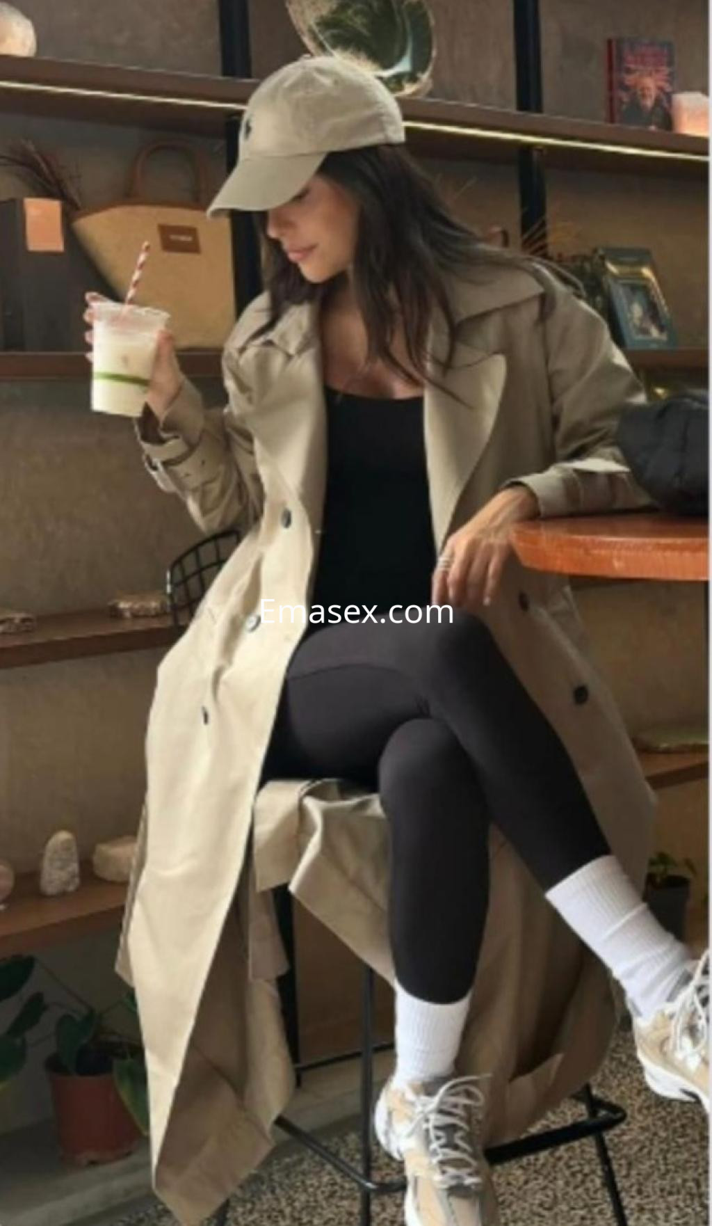 Puta en Bilbao sentada en una cafetería, vistiendo un abrigo beige, leggings oscuros y zapatillas deportivas.