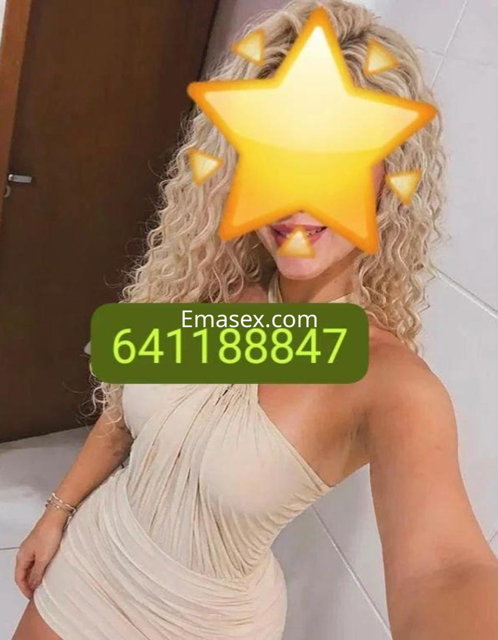 Puta con cabello rizado y rubio, viste un vestido ajustado color beige, posando en un entorno luminoso en Alicante.