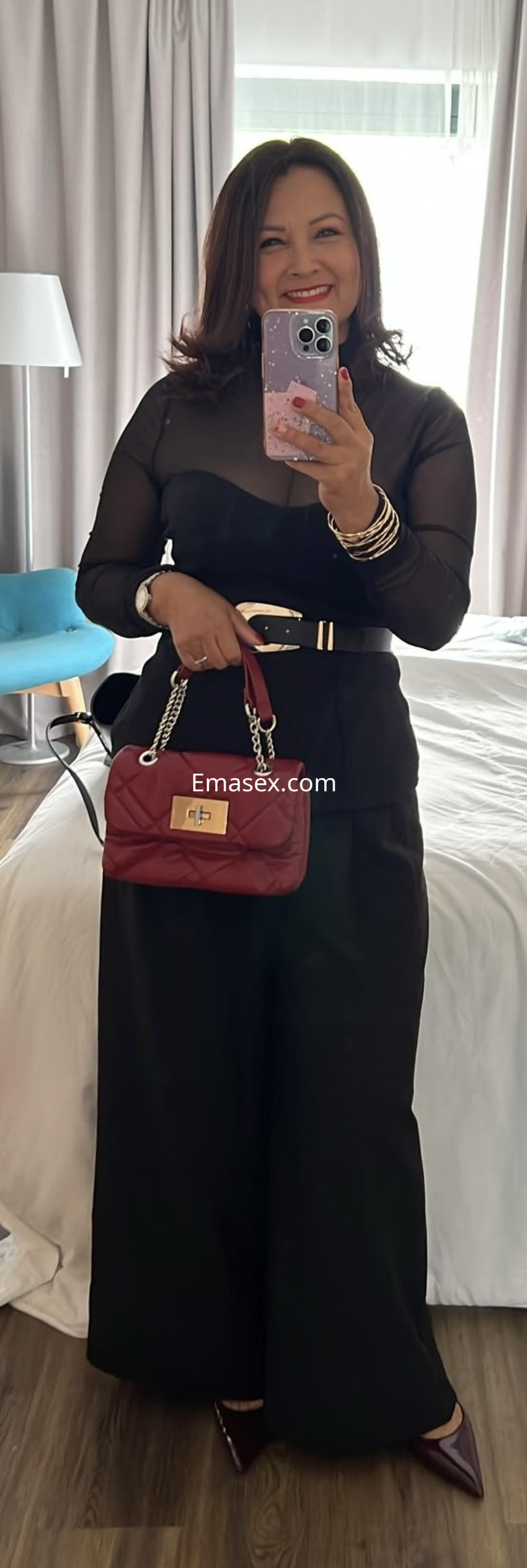 Puta con cabello oscuro y blusa transparente, posando con un bolso rojo en un dormitorio moderno en Santander.