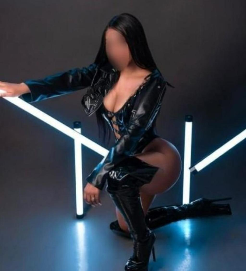 Escort en Madrid, de piel morena, viste un atuendo de cuero negro y tacones altos, posando de rodillas con luces de fondo.