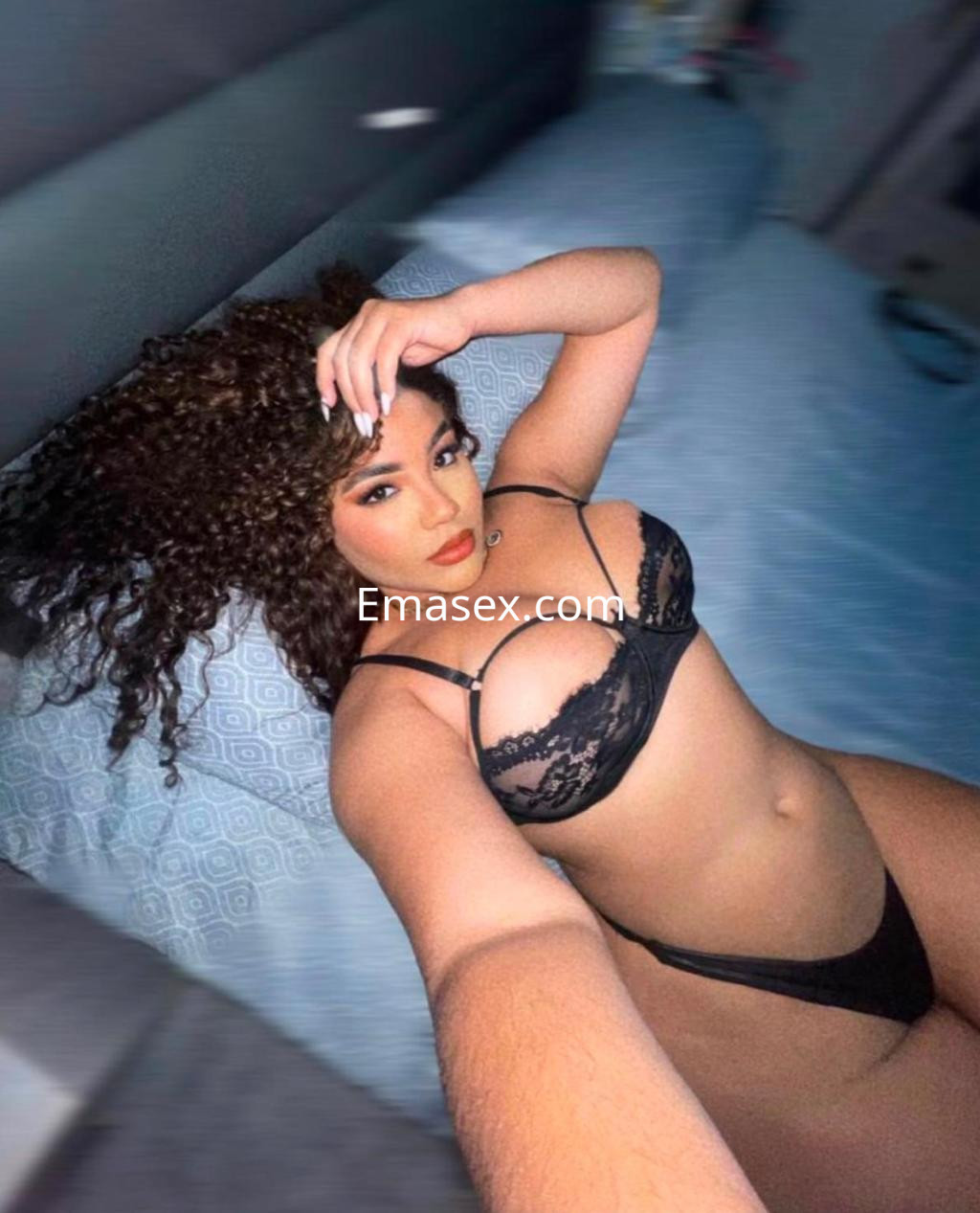 Escort en Vitoria posando en la cama con lencería negra, cabello rizado y expresión seductora.