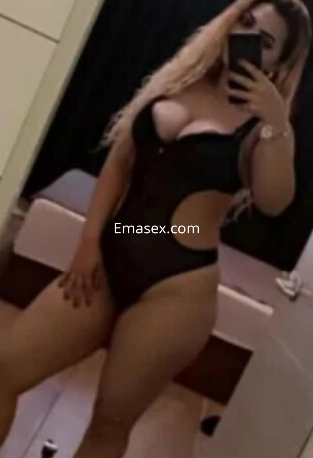 Escort en Gijón vistiendo un body negro ajustado, posando frente a un espejo con cabello rubio ondulado.