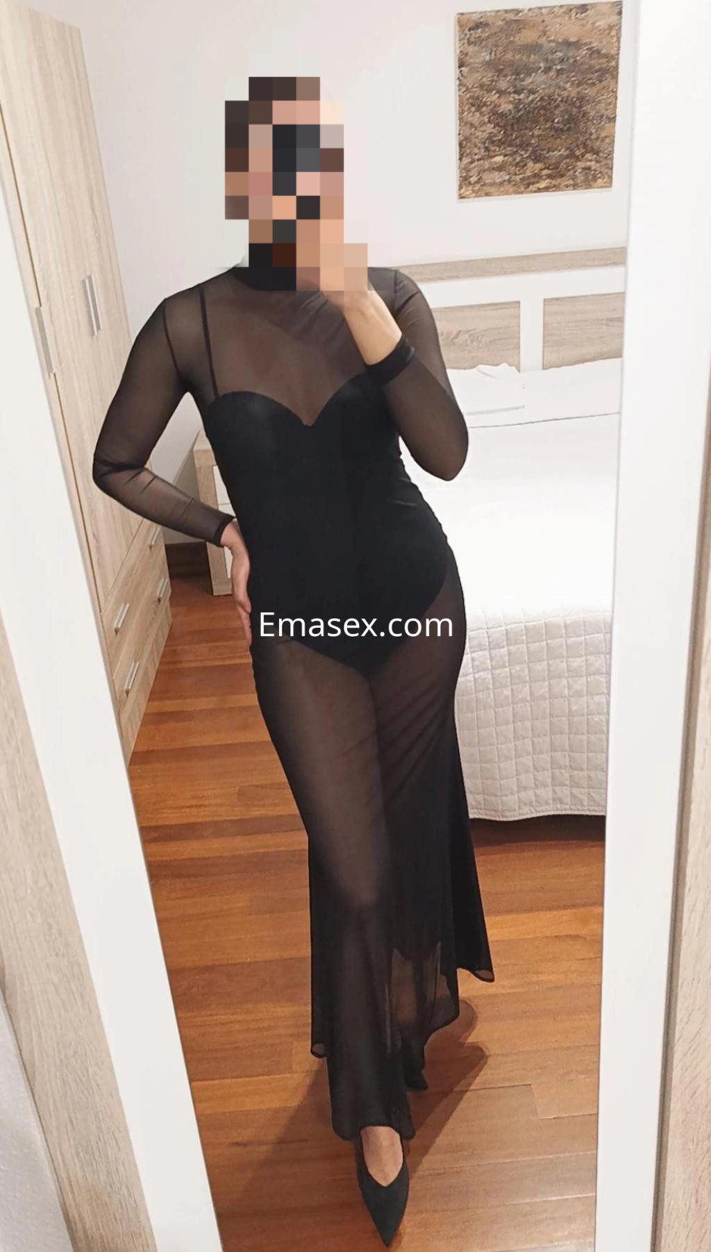 Puta en Santander posando frente a un espejo con un vestido negro transparente de manga larga y tacones.