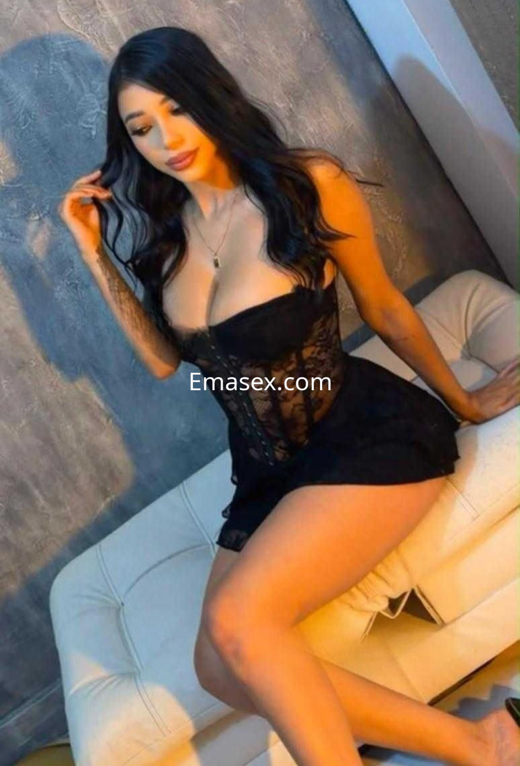 Puta en Alicante con cabello ondulado y oscuro, viste un vestido corto negro y posa sentada en un sofá claro.