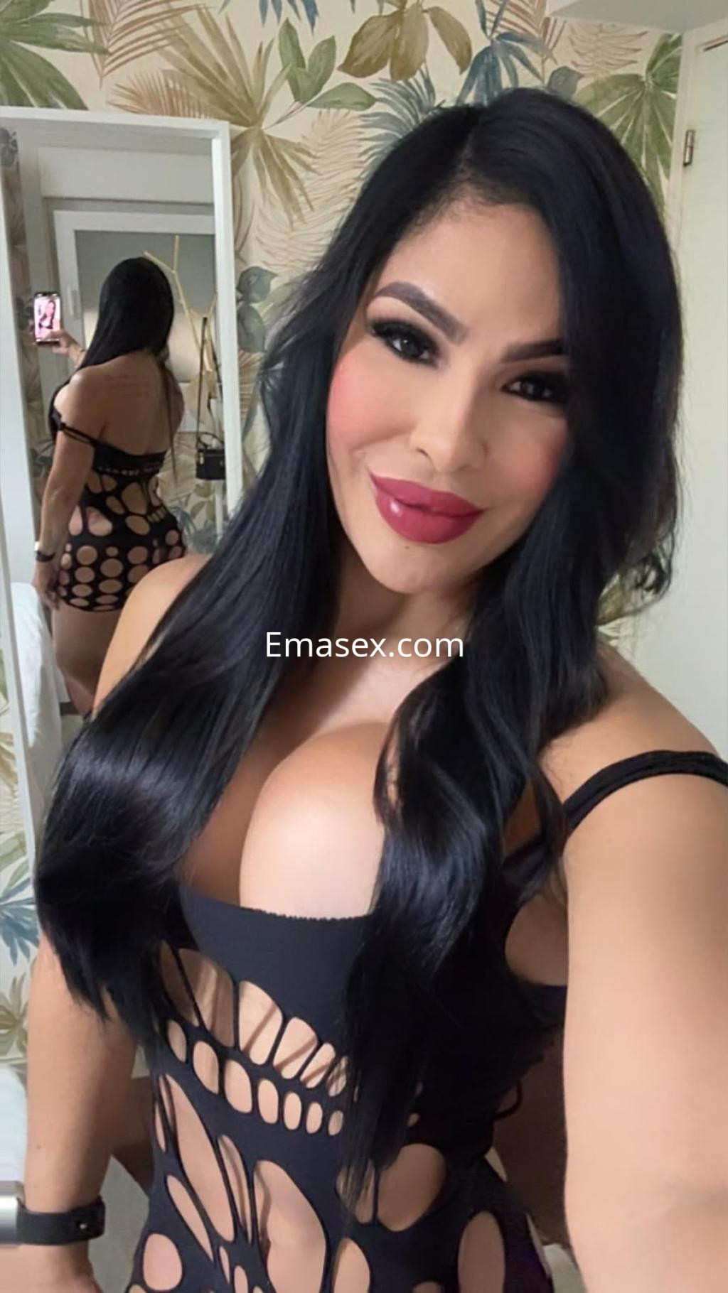 Puta tomando una selfie en un ambiente decorado con hojas, luciendo un vestido negro con cortes y cabello largo.