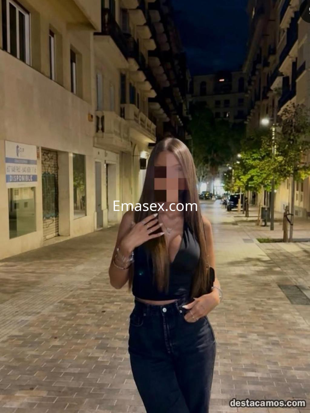 Escort en León, posando en la calle de noche con un top negro y jeans ajustados, sonriendo ligeramente.