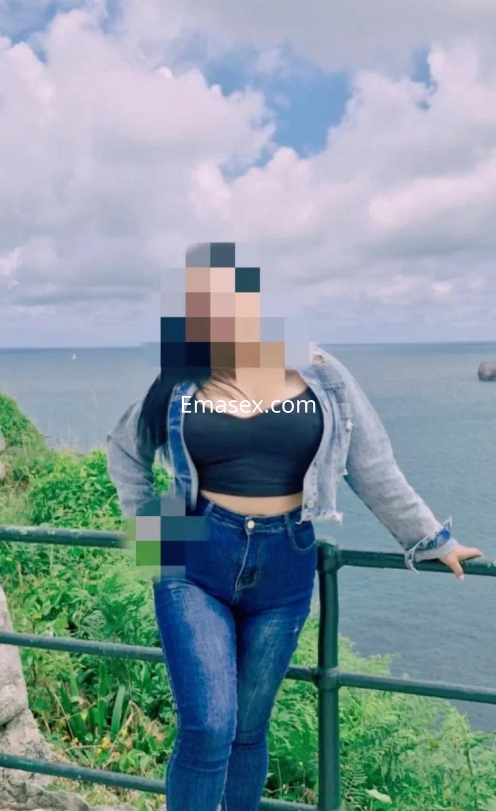 Escort en Murcia viste un top negro y jeans ajustados, posando junto al mar con un cielo nublado.