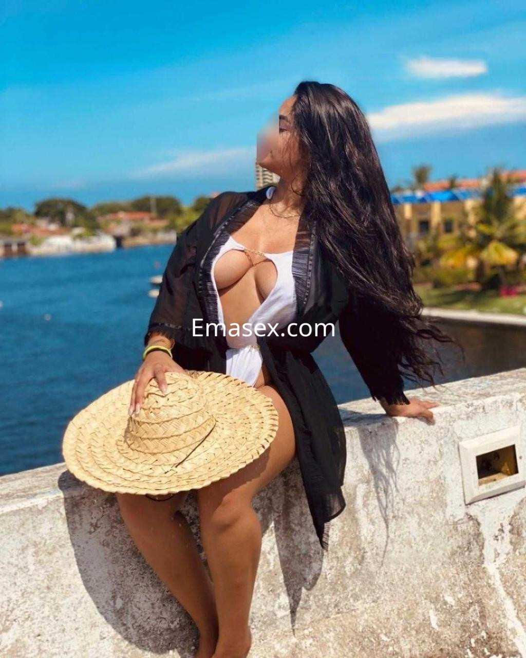 Puta posando en Alicante con un traje de baño blanco, un sombrero de paja y un fondo de agua azul.
