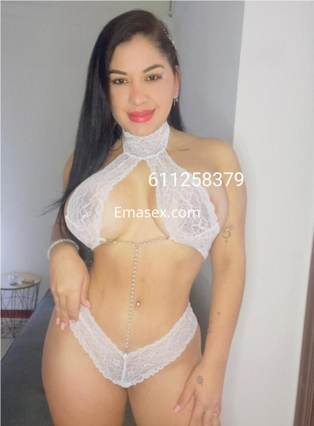Escort en Almería, posando con lencería blanca de encaje y un collar, en un ambiente iluminado y minimalista.