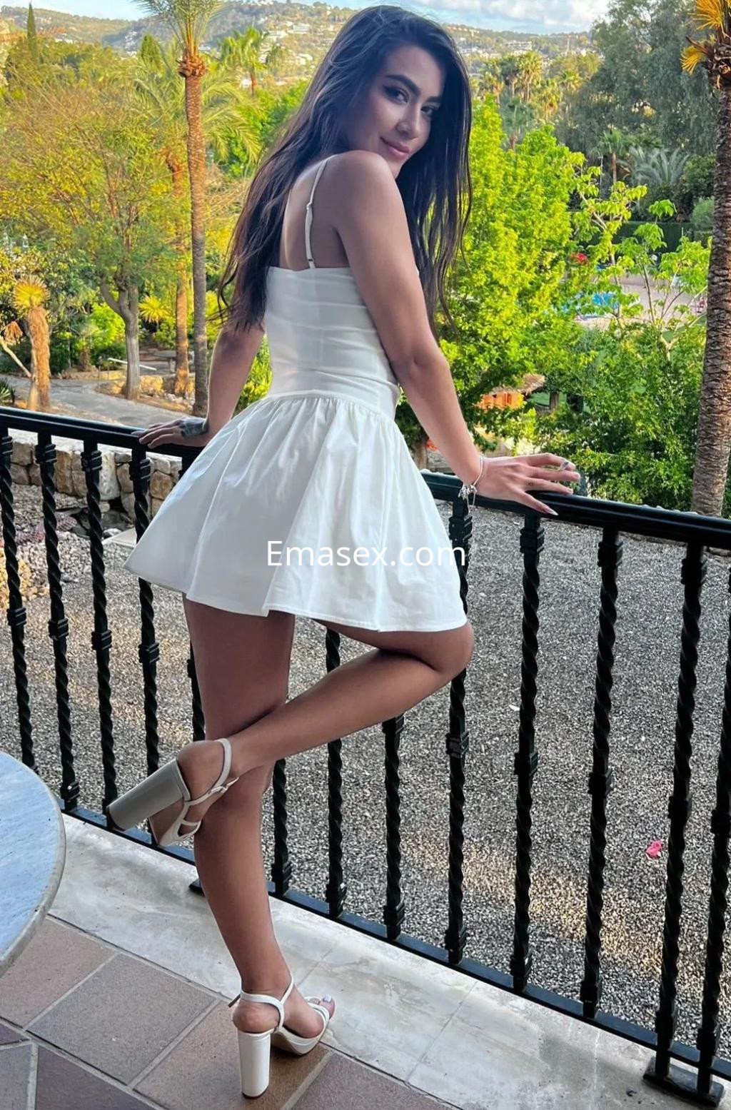 La puta luce un vestido blanco corto y sandalias altas, posando con confianza en un entorno natural en Palma de Mallorca.