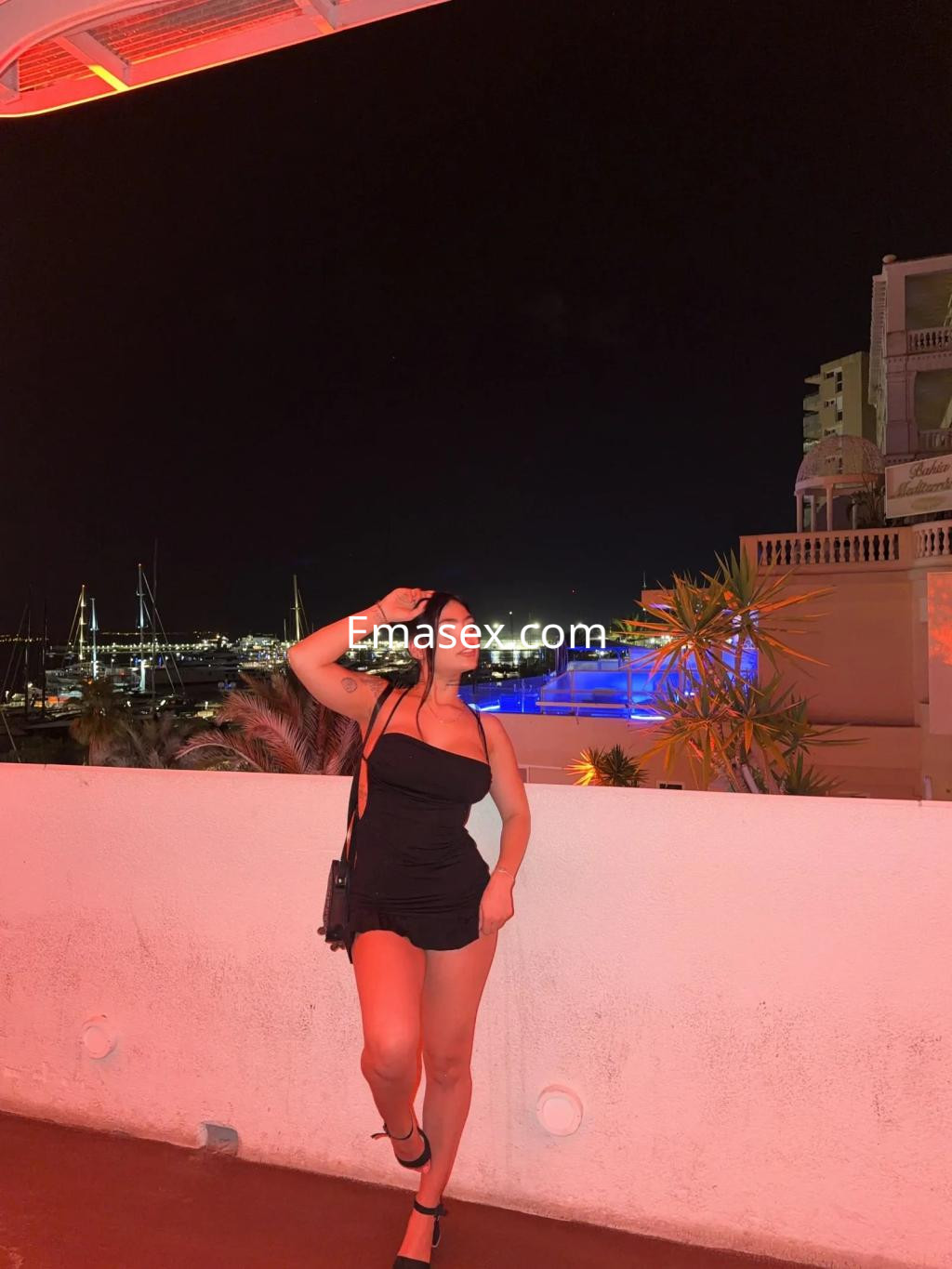 Escort posando en un entorno nocturno en Palma de Mallorca, vestida con un vestido negro ajustado y sandalias.