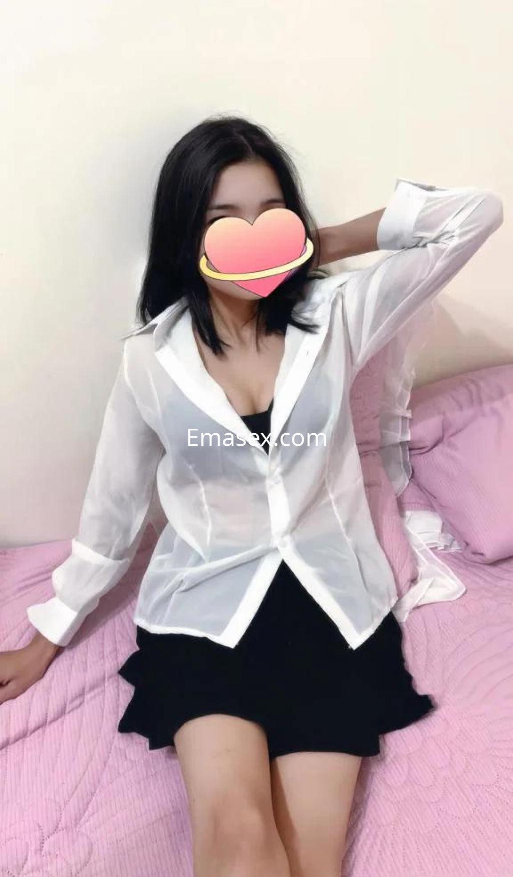 Escort en Barcelona sentada en una cama, luciendo una blusa blanca semi transparente y una falda negra.