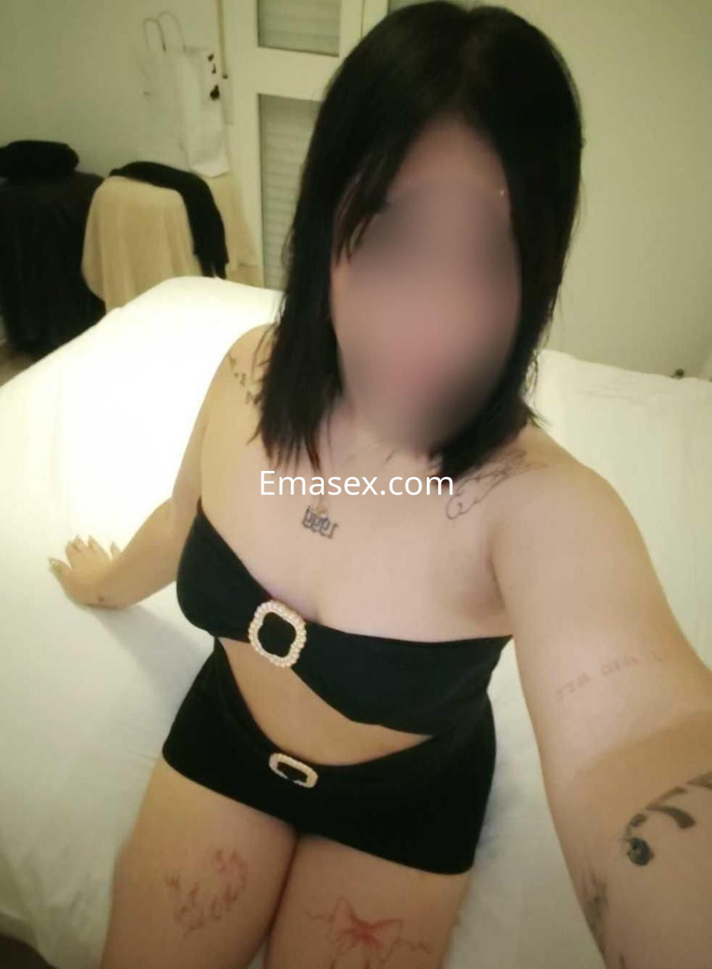 Escort en Cádiz posando en una cama, lleva un conjunto negro con detalles de cinturón y muestra varios tatuajes.