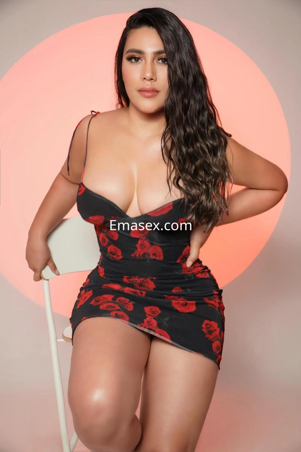 Escort en Cádiz, luciendo un vestido ajustado negro con estampado de rosas rojas, posando sentada en una silla.