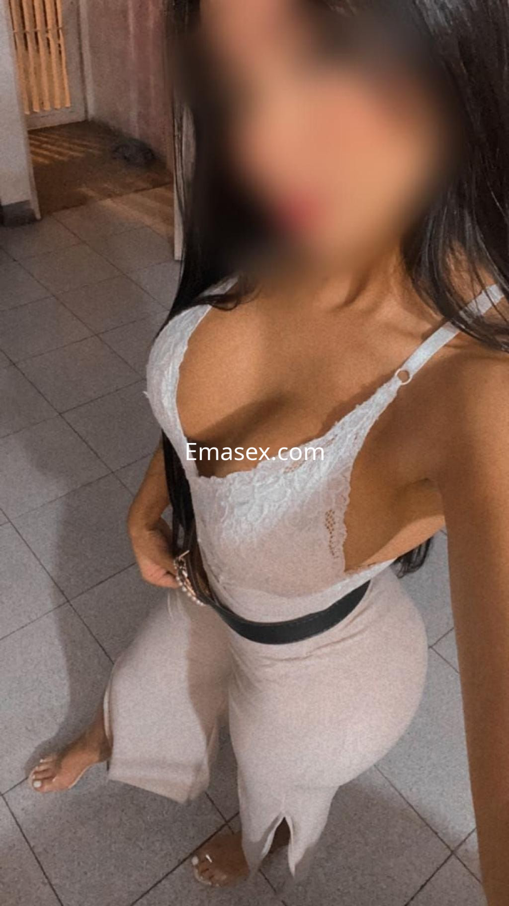 Escort en Coruña posando en un ambiente interior, vistiendo un conjunto claro con detalles de encaje y cinturón negro.