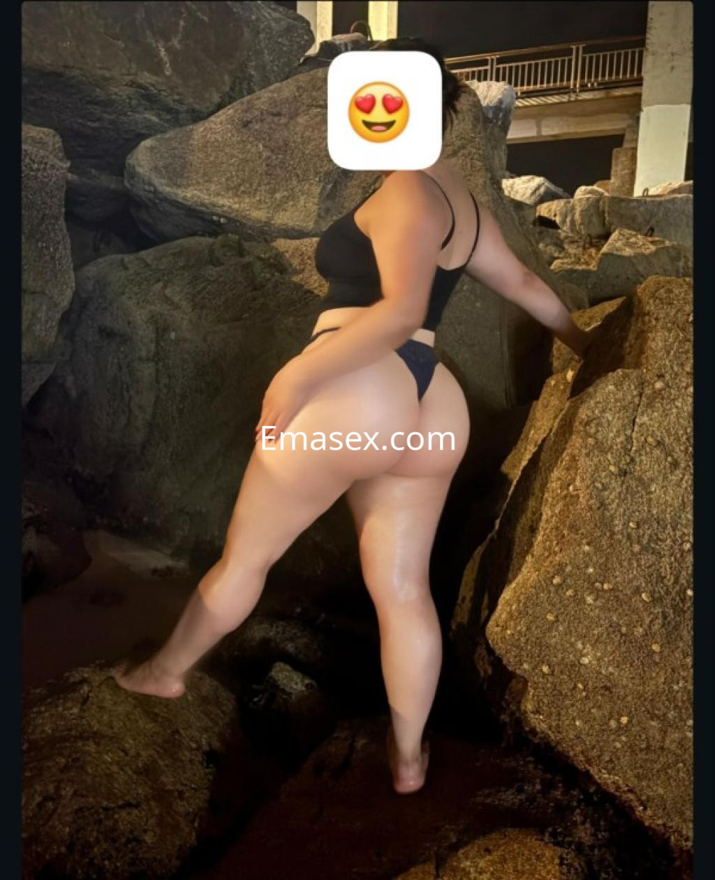 Escort en Coruña posando de espaldas sobre rocas, vistiendo un bikini negro que resalta su figura.