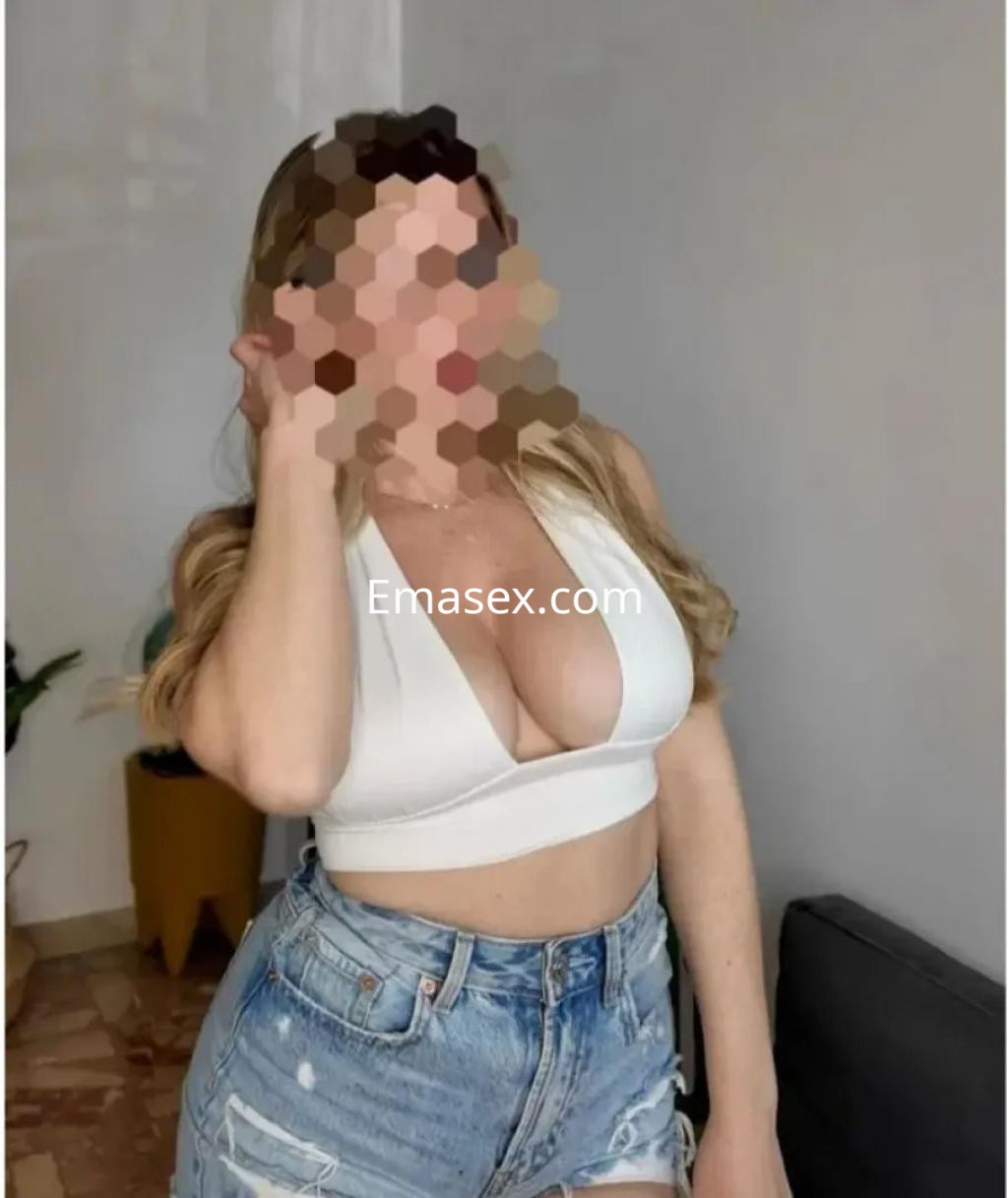 Escort en Murcia con cabello largo y ondulado, vistiendo un top blanco y shorts de mezclilla, sonriente.
