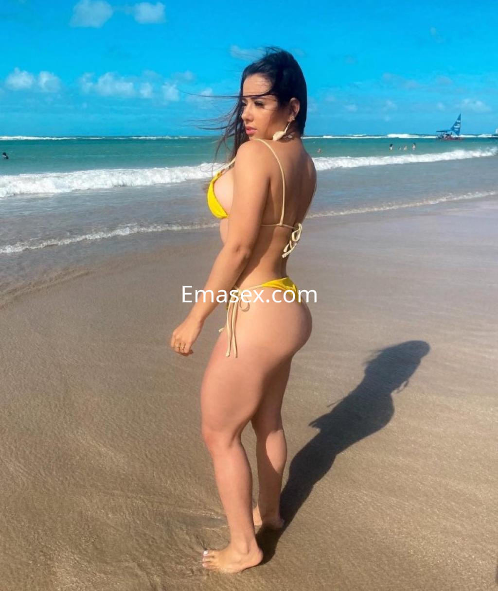 Escort en Tarragona posando de espaldas en la playa, con bikini amarillo y el mar de fondo.
