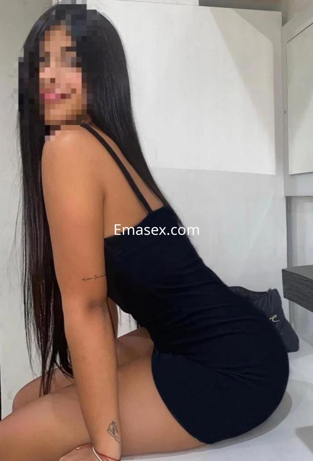 La puta en Valladolid lleva un vestido negro ajustado, sentada en una superficie plana con fondo claro.