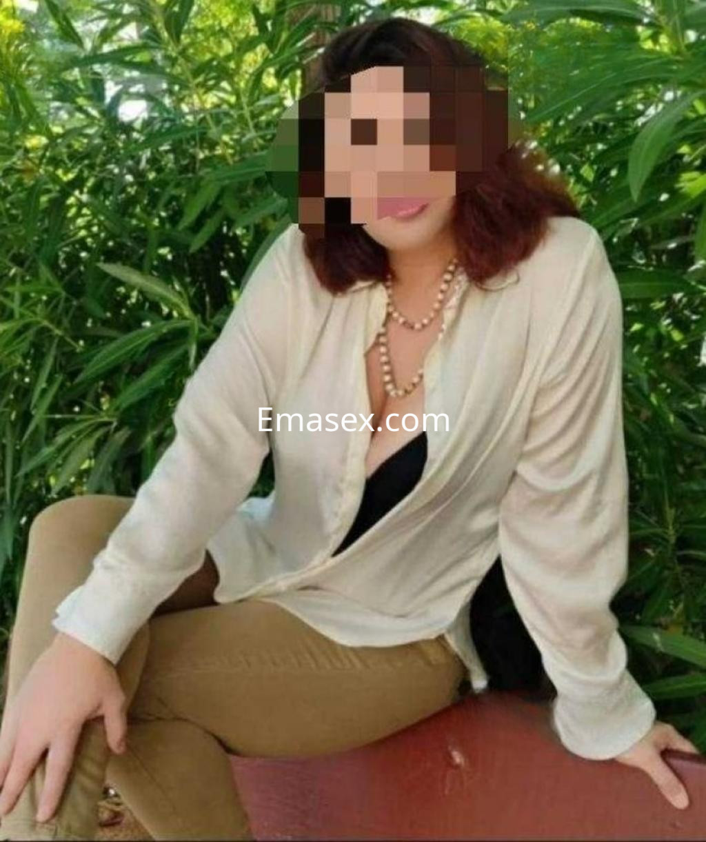 La escort en Madrid luce una blusa clara con un escote pronunciado y pantalones ajustados, rodeada de vegetación.