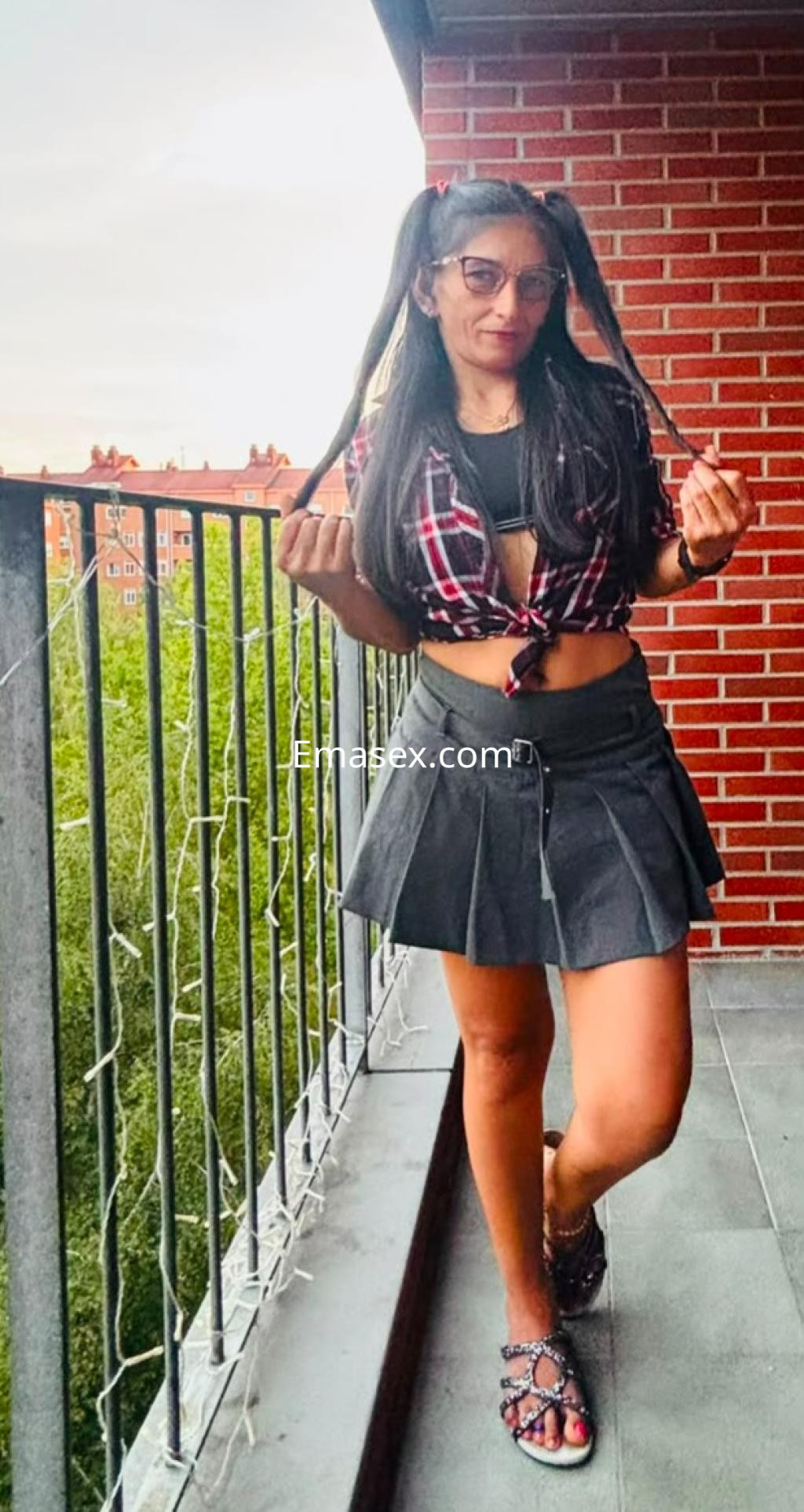 Escort en Vitoria, luce una blusa a cuadros y una falda corta, posando en un balcón con vista a la ciudad.