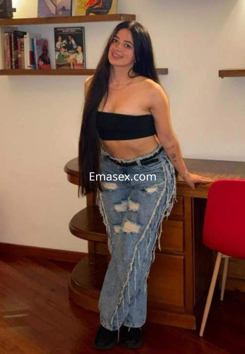 Escort en Madrid con cabello largo y oscuro, viste un top negro y jeans desgastados, posando junto a un mueble.