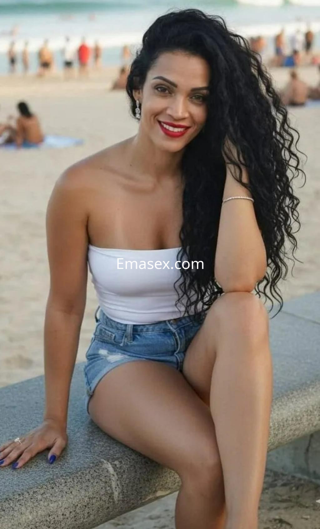 Escort en Madrid con cabello rizado y oscuro, vestida con top blanco y shorts de mezclilla, sonriendo en la playa.