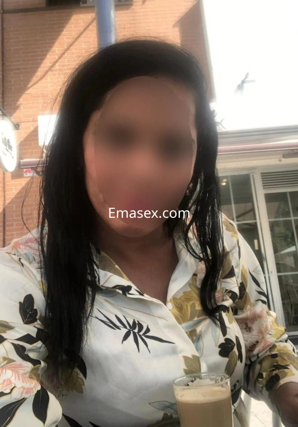 Escort en Valladolid con cabello largo y oscuro, usando una blusa floral mientras sostiene una bebida en un ambiente exterior.