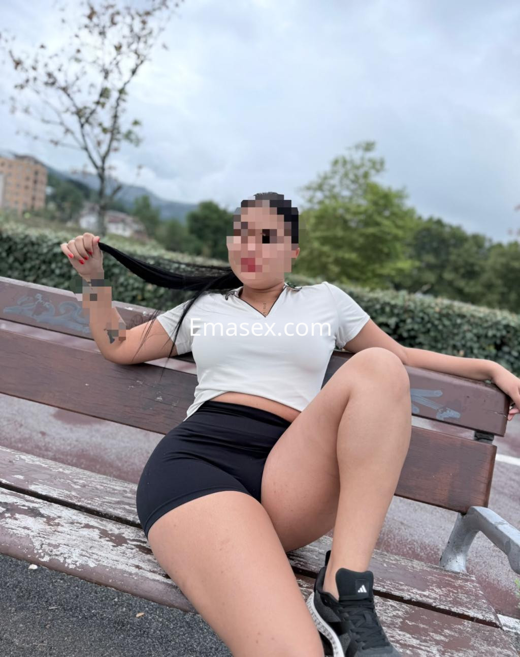 Escort sentada en un banco en Irún, vistiendo una camiseta blanca y shorts negros, con cabello largo y lacio.