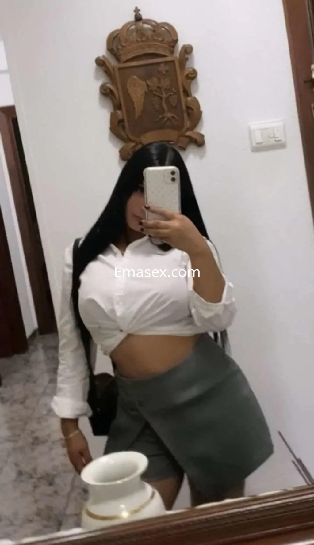 Escort en Coruña, con blusa blanca y falda corta gris, posando frente a un espejo en un ambiente interior.