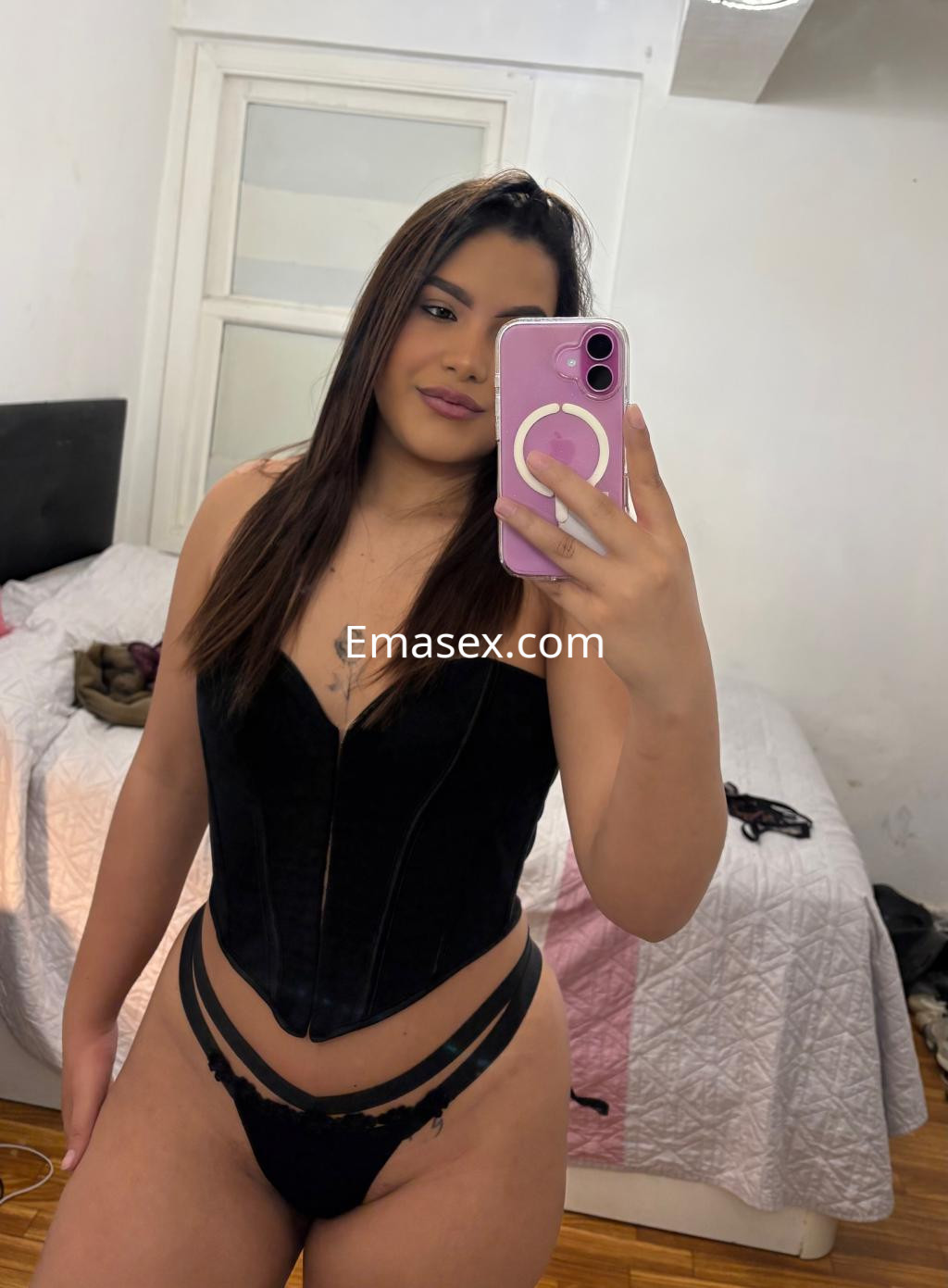 Puta en Vigo con cabello largo y lacio, vistiendo un corset negro y tanga, posando en un dormitorio con cama.