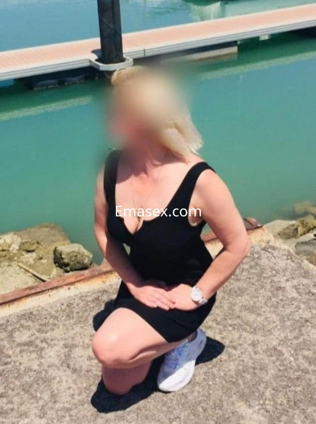 Escort en Málaga, de cabello rubio y piel clara, viste un vestido negro y se arrodilla junto al agua.
