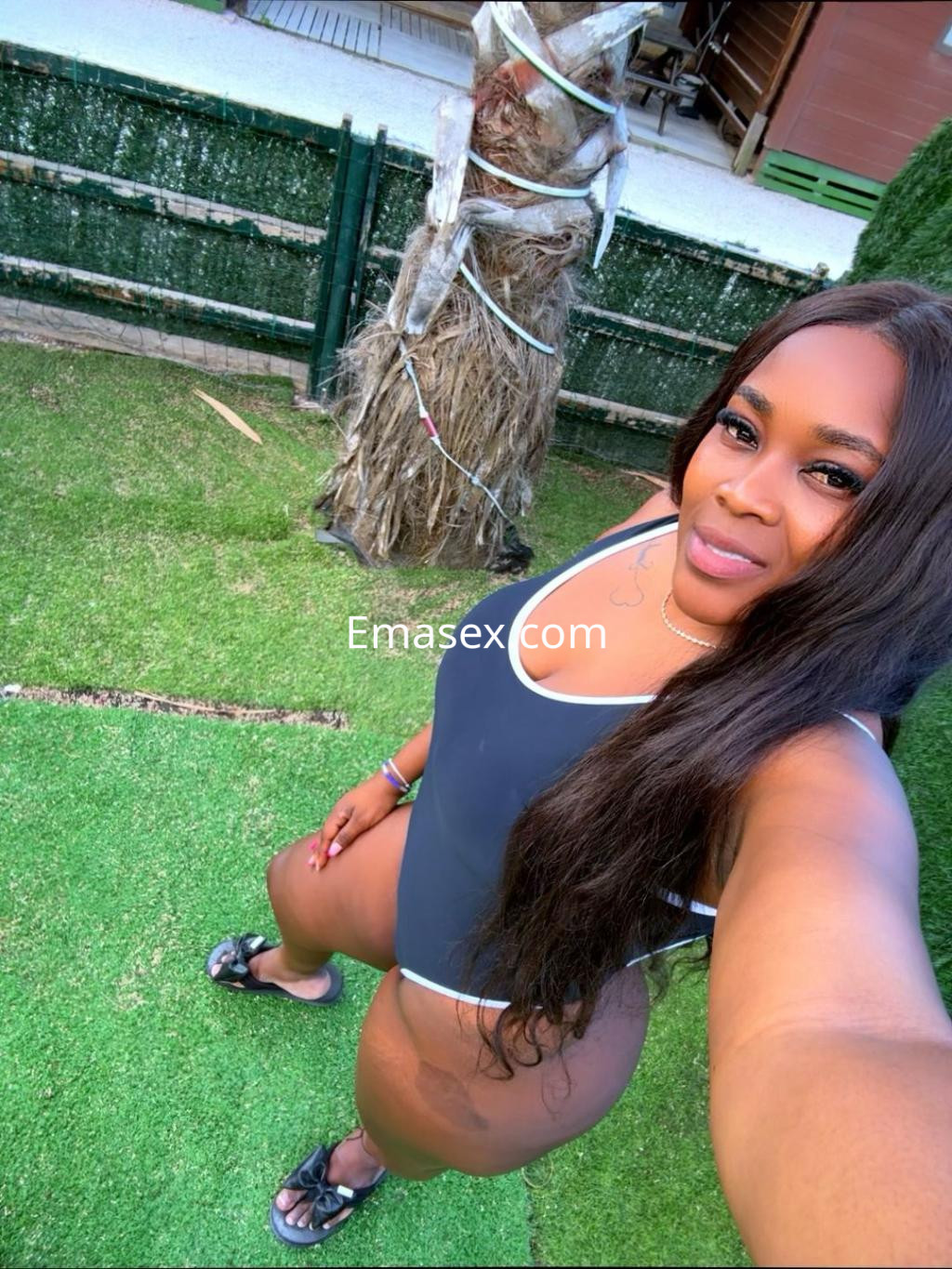 Escort en Irún, posando en un jardín con un traje de baño negro y sandalias, luciendo una sonrisa y cabello largo.