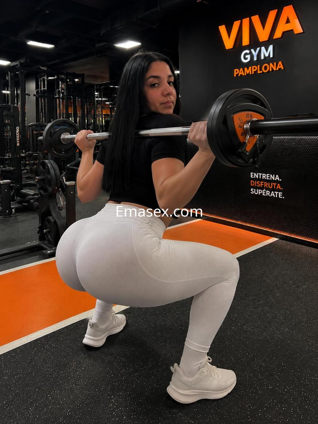 Escort en Pamplona, con mallas blancas y camiseta negra, levantando pesas en un gimnasio moderno.