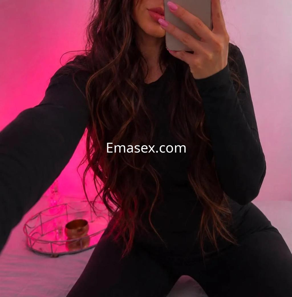 Puta en Valencia con cabello largo y ondulado, vestida de negro, tomando una selfie en un ambiente iluminado.
