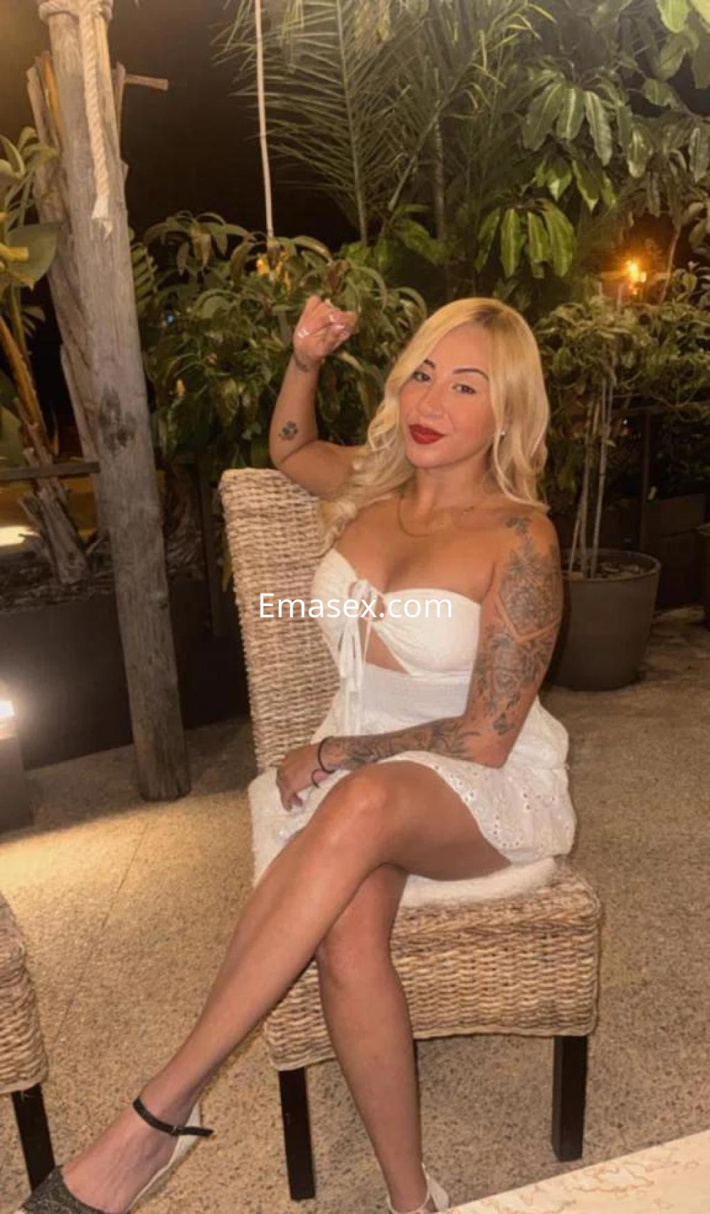 Escort en Castellón sentada en una silla, luciendo blusa blanca sin mangas y falda corta, con cabello rubio y tatuajes.