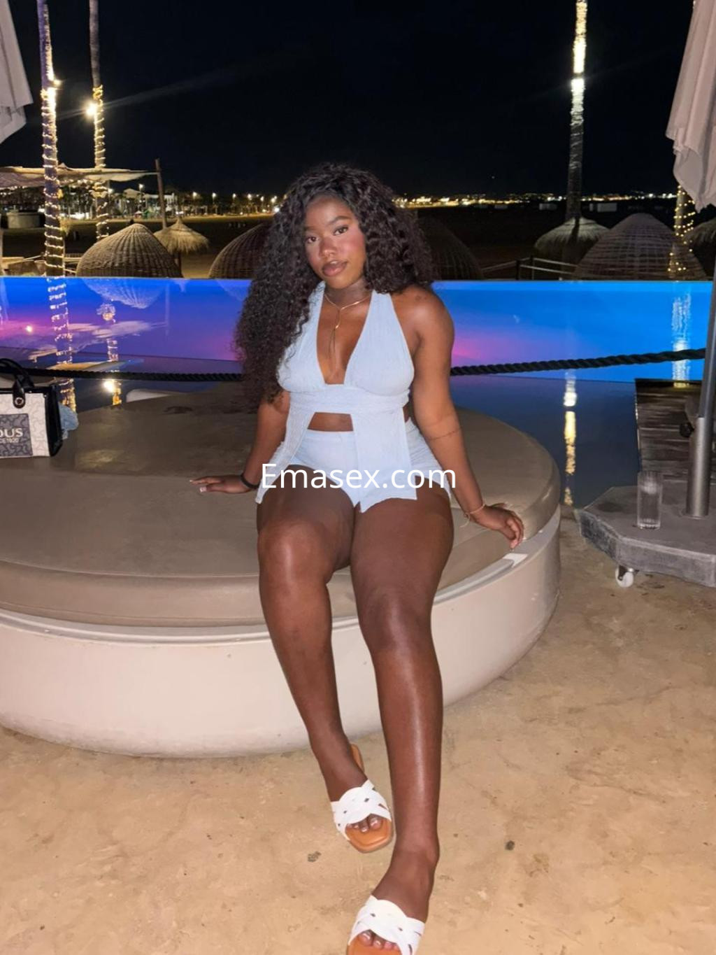 La escort se encuentra sentada en un entorno nocturno en Valencia, vistiendo un conjunto azul claro y sandalias blancas.