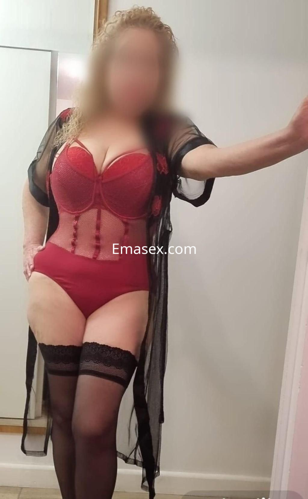 La puta luce un conjunto de lencería rojo con medias negras, posando en un entorno interior en Cádiz.