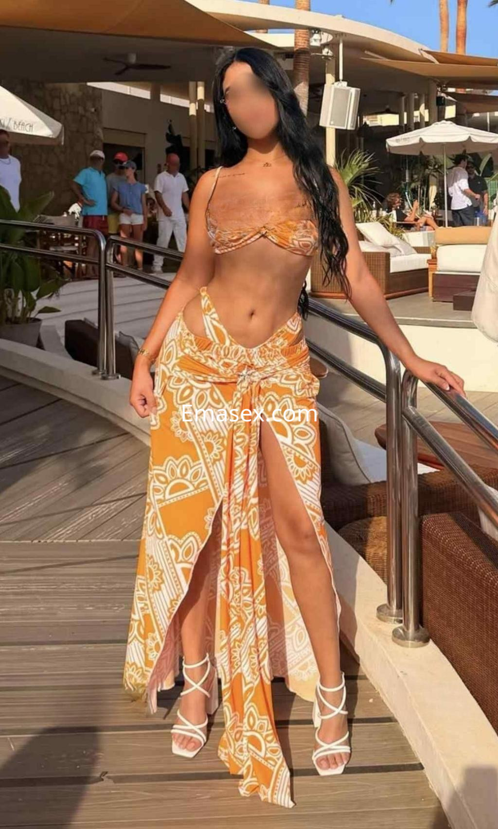 Escort en Madrid, con cabello largo y oscuro, viste un bikini naranja y una falda larga con estampado, posando al aire libre.