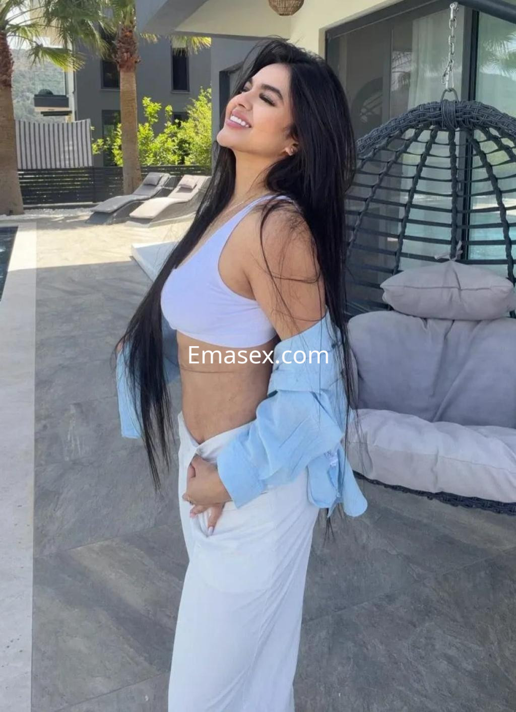 Escort en Madrid, de pie en un entorno exterior, con cabello largo y una blusa azul claro, luciendo una sonrisa.